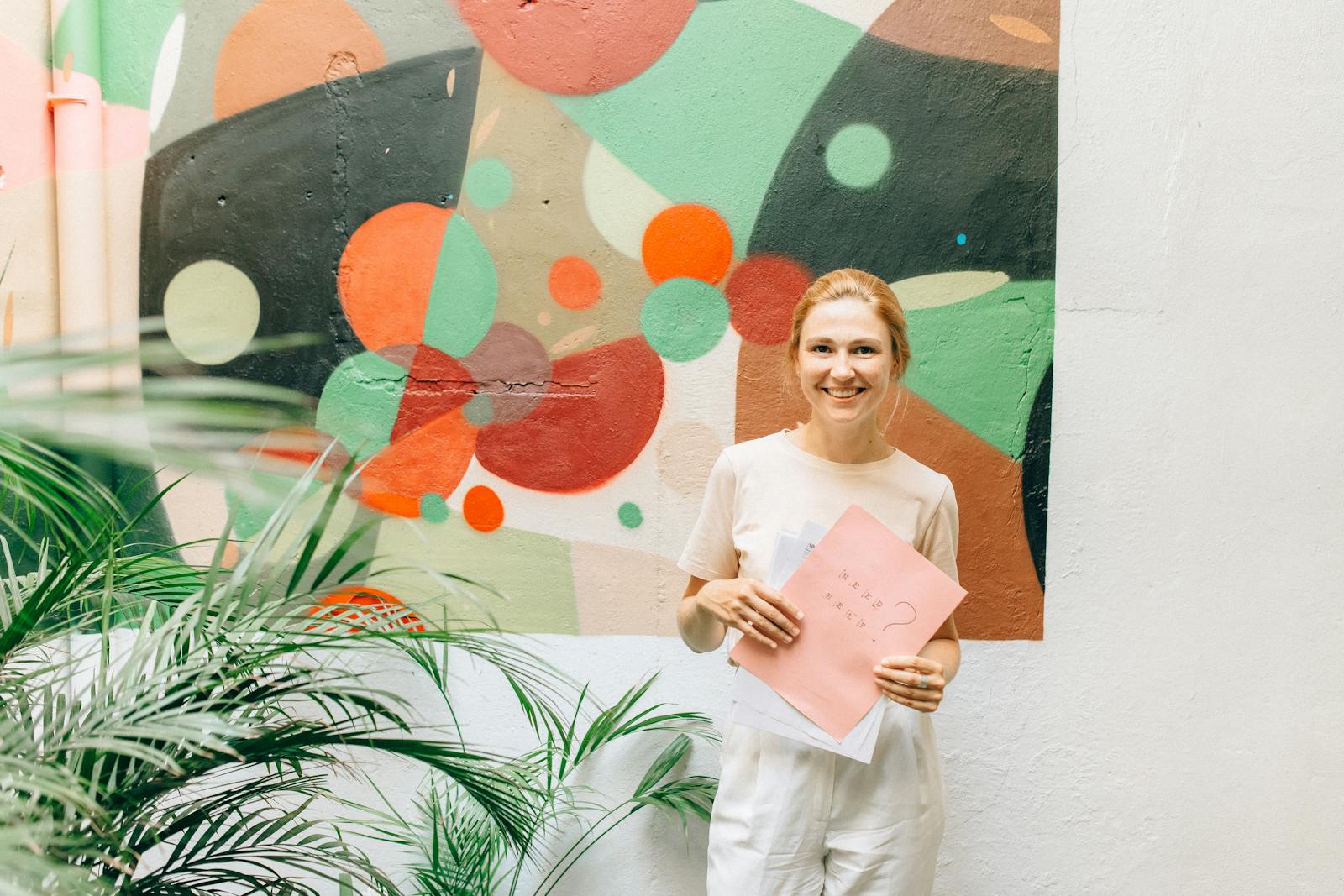 A cheerful woman in a white outfit holds papers in front of a vibrant abstract mural.